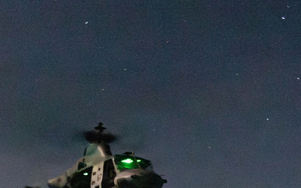 A military helicopter, appearing to be a UH-1Y Venom or AH-1Z Viper type, is captured lifting off or hovering low over a naval vessel's flight deck at night. The aircraft displays bright green navigation/IR lights and bears a visible side number. A single deck crew member in flight gear stands on the darkened flight deck observing the aircraft.