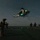 LCF-24: Lowlight Fast-roping drills A nighttime military helicopter operation taking place on what appears to be a naval vessel's flight deck. A helicopter, likely a UH-72 Lakota or similar utility helicopter, hovers low above the deck with green navigation lights illuminated while connected to personnel below via a rope or cable. Multiple armed and equipped military personnel are visible on the deck participating in what appears to be a fast-rope or hoist training exercise.