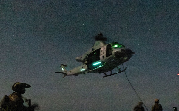 A nighttime military helicopter operation taking place on what appears to be a naval vessel's flight deck. A helicopter, likely a UH-72 Lakota or similar utility helicopter, hovers low above the deck with green navigation lights illuminated while connected to personnel below via a rope or cable. Multiple armed and equipped military personnel are visible on the deck participating in what appears to be a fast-rope or hoist training exercise.