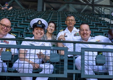 A group of U.S. Navy personnel in white dress uniforms are seated in stadium bleachers, posing and smiling for the camera. The group includes officers of various ranks, identifiable by their rank insignia, ribbons, and cover (hat), along with at least one civilian. They appear to be attending a sporting event in a large stadium.