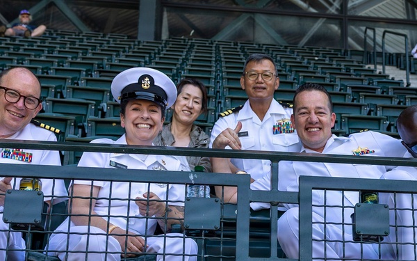 A group of U.S. Navy personnel in white dress uniforms are seated in stadium bleachers, posing and smiling for the camera. The group includes officers of various ranks, identifiable by their rank insignia, ribbons, and cover (hat), along with at least one civilian. They appear to be attending a sporting event in a large stadium.