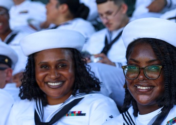 A group of U.S. Navy enlisted personnel in white dress uniforms (Service Dress White) are seated together, likely at a public event or ceremony. Two female sailors in the foreground are smiling and posing for the photo, both wearing white Dixie cup covers and black neckerchiefs with service ribbons visible. A male officer in a white combination cover with gold insignia is visible to the left.