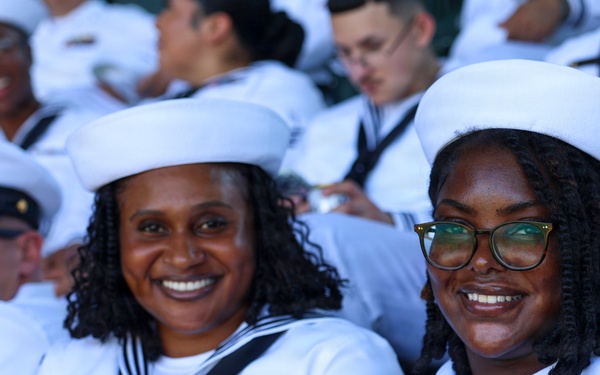 A group of U.S. Navy enlisted personnel in white dress uniforms (Service Dress White) are seated together, likely at a public event or ceremony. Two female sailors in the foreground are smiling and posing for the photo, both wearing white Dixie cup covers and black neckerchiefs with service ribbons visible. A male officer in a white combination cover with gold insignia is visible to the left.