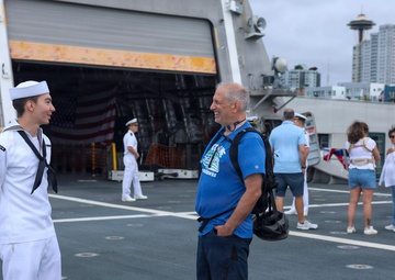 A U.S. Navy sailor in dress white uniform converses with a civilian visitor on the deck of a naval vessel, likely during a public open-ship event. The Seattle Space Needle is visible in the background, indicating this is taking place in Seattle, Washington. The scene appears to be a Fleet Week or similar public outreach event.