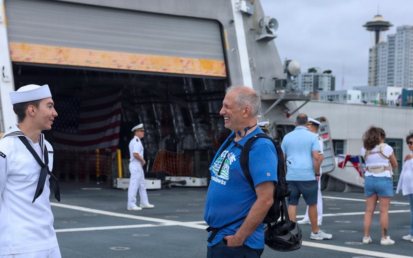 A U.S. Navy sailor in dress white uniform converses with a civilian visitor on the deck of a naval vessel, likely during a public open-ship event. The Seattle Space Needle is visible in the background, indicating this is taking place in Seattle, Washington. The scene appears to be a Fleet Week or similar public outreach event.