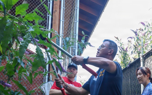 Three people are working in an outdoor garden area alongside a chain-link fence. A man in a navy blue 'NAVY' shirt is using a long-handled tool, possibly loppers or pruning shears, to trim overhead branches, while a man in a red shirt assists by holding the tool or branches. A woman in a plaid shirt observes nearby.
