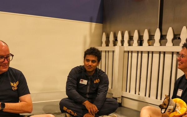 Three people are seated on the floor in what appears to be an indoor facility, two of whom are holding small puppies wearing protective cone collars. All three individuals are wearing matching dark navy t-shirts with a gold eagle logo, suggesting they are part of the same organization or team. The setting includes a white picket fence backdrop and a gray blanket on the floor.