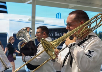 Two U.S. Navy musicians in dress white uniforms perform outdoors, with one playing a trombone and another playing a euphonium or baritone horn. A music stand with sheet music is visible in the foreground. The setting appears to be a waterfront or dock area, likely during a public event or Navy Week performance.