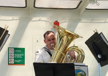 A U.S. Navy musician in white dress uniform plays a tuba on what appears to be the deck of a vessel. A music stand in front of him displays a label reading 'Tuba' along with a logo referencing 'Band Northwest' and 'Silverdale, Washington' and 'Brass Quintet'. The setting includes safety signage and other music stands visible in the background.