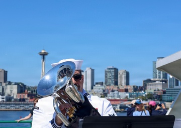 A U.S. Navy musician in dress whites plays a euphonium or baritone horn on a waterfront pier in Seattle, Washington, identifiable by the Space Needle visible in the background skyline. A music stand in the foreground displays what appears to be a Navy band insignia, partially readable as 'Northwest Washington' and 'Quintet'. The scene appears to be a public outdoor performance event on a clear, sunny day.