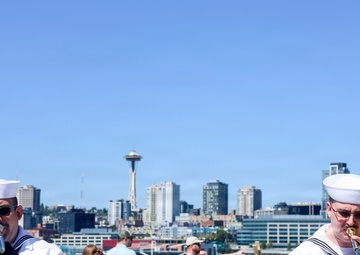 Two U.S. Navy musicians in dress white uniforms are performing outdoors on what appears to be a waterfront deck or vessel, with the Seattle skyline and Space Needle clearly visible in the background. They are playing brass instruments — a mellophone or marching French horn on the left and a trombone on the right — with music stands bearing what appears to be a Navy band insignia. The scene takes place on a clear, sunny day with civilians visible in the background.