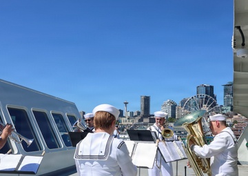 Members of the Navy Band Northwest, based in Silverdale, Washington, perform an outdoor brass ensemble concert on the deck of a naval vessel. The musicians, dressed in white Navy dress uniforms, play various brass instruments with music stands in front of them. The Seattle skyline, including the Space Needle and a Ferris wheel, is visible in the background.