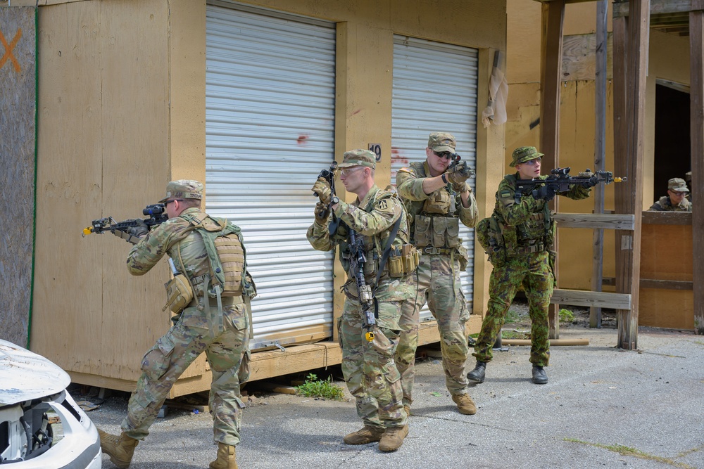 Four soldiers in military uniforms are conducting what appears to be a tactical training exercise in an urban operations environment. Three soldiers wear US Army Operational Camouflage Pattern (OCP) uniforms while one wears a distinctive green woodland camouflage pattern consistent with a foreign military. They are moving in formation with rifles raised in a clearing or building-entry drill scenario.