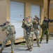 Four soldiers in military uniforms are conducting what appears to be a tactical training exercise in an urban operations environment. Three soldiers wear US Army Operational Camouflage Pattern (OCP) uniforms while one wears a distinctive green woodland camouflage pattern consistent with a foreign military. They are moving in formation with rifles raised in a clearing or building-entry drill scenario.