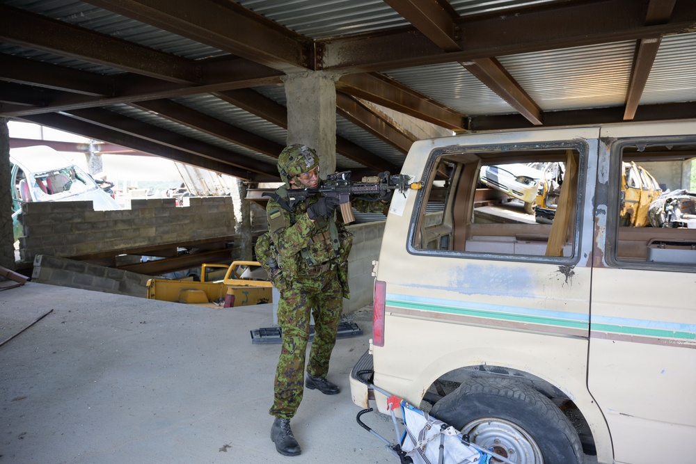 A soldier in camouflage uniform and helmet is aiming a rifle inside what appears to be an industrial or warehouse structure. The setting resembles a military training exercise or urban operations drill, with a dilapidated van and scattered debris visible in the background. The environment appears to be a mock urban combat training area.
