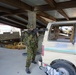 A soldier in camouflage uniform and helmet is aiming a rifle inside what appears to be an industrial or warehouse structure. The setting resembles a military training exercise or urban operations drill, with a dilapidated van and scattered debris visible in the background. The environment appears to be a mock urban combat training area.