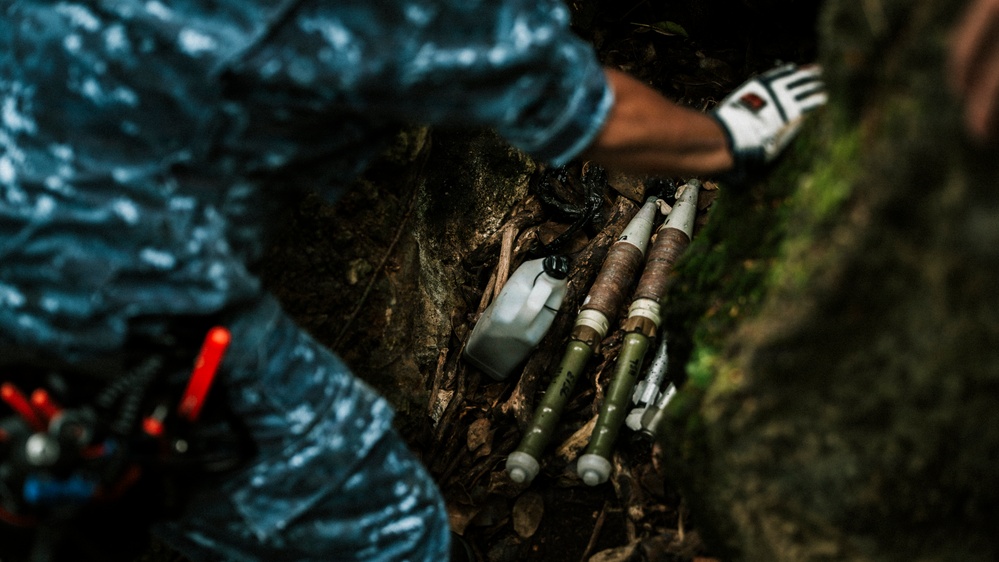 A person wearing a blue uniform and tactical gloves is examining or handling what appear to be mortar rounds or rocket-propelled munitions found in a concealed outdoor location among rocks and leaf litter. The munitions appear to be cached or hidden in a natural crevice. The scene suggests a military or law enforcement discovery/recovery operation.