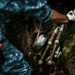 A person wearing a blue uniform and tactical gloves is examining or handling what appear to be mortar rounds or rocket-propelled munitions found in a concealed outdoor location among rocks and leaf litter. The munitions appear to be cached or hidden in a natural crevice. The scene suggests a military or law enforcement discovery/recovery operation.