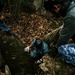 An overhead view of military or law enforcement personnel in digital camouflage uniforms navigating rocky terrain in a forested environment. The central figure wears a black helmet and white tactical gloves, positioned between large moss-covered boulders. Several cylindrical objects, possibly ordnance or equipment, are visible at the bottom of the rocky crevice.