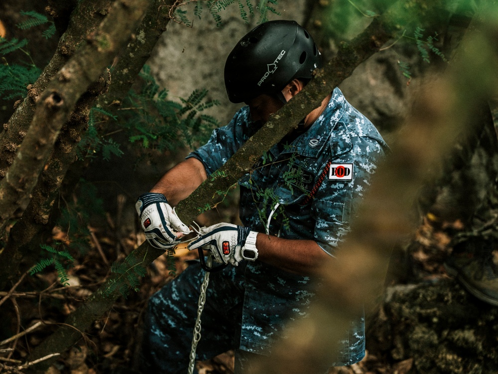 A person wearing a black Pro-Tec helmet, blue digital camouflage shirt, and white gloves is working in a forested environment. They appear to be handling a rope or chain attached to a tree branch or log. The setting is dense woodland with visible rocks, ferns, and fallen leaves.