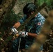 A person wearing a black Pro-Tec helmet, blue digital camouflage shirt, and white gloves is working in a forested environment. They appear to be handling a rope or chain attached to a tree branch or log. The setting is dense woodland with visible rocks, ferns, and fallen leaves.