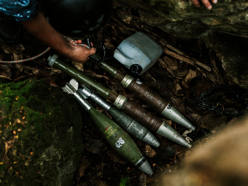 The image shows multiple mortar rounds or artillery shells laid out on a rocky, leaf-covered ground in an outdoor setting. A person's hand is visible at the top of the frame, appearing to handle or inspect the munitions. The ordnance appears to be of military origin, olive-drab in color with visible markings.
