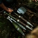 The image shows multiple mortar rounds or artillery shells laid out on a rocky, leaf-covered ground in an outdoor setting. A person's hand is visible at the top of the frame, appearing to handle or inspect the munitions. The ordnance appears to be of military origin, olive-drab in color with visible markings.