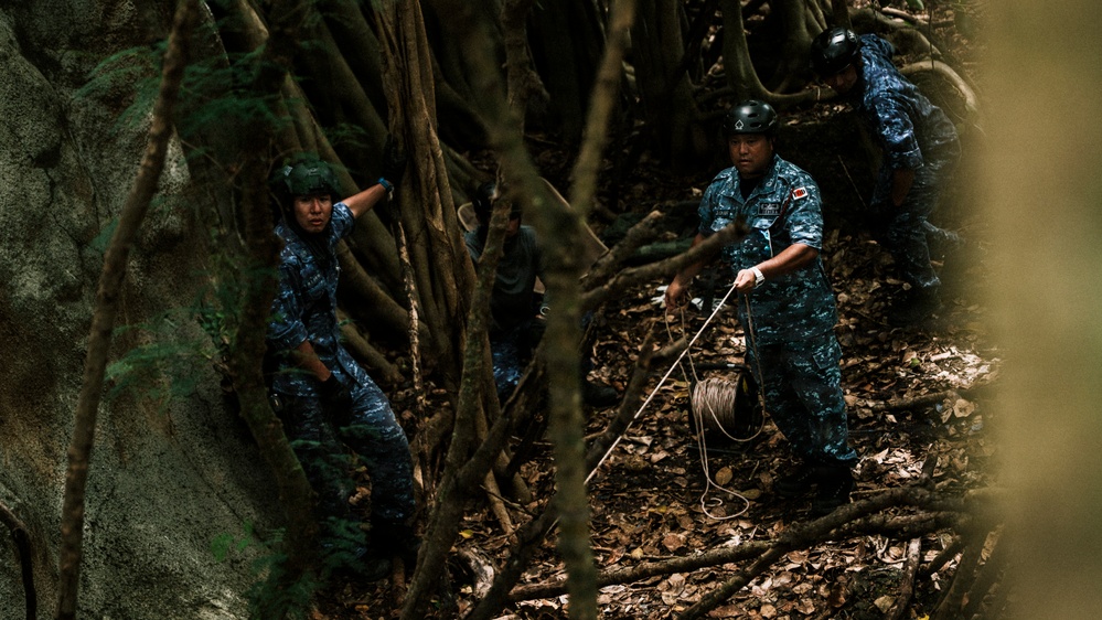 Military or law enforcement personnel in blue digital camouflage uniforms and helmets are navigating through dense jungle terrain with large tree roots. One individual is visibly carrying a coil of rope or cable. The setting appears to be a training exercise or operational mission in a tropical forest environment.