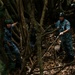 Military or law enforcement personnel in blue digital camouflage uniforms and helmets are navigating through dense jungle terrain with large tree roots. One individual is visibly carrying a coil of rope or cable. The setting appears to be a training exercise or operational mission in a tropical forest environment.