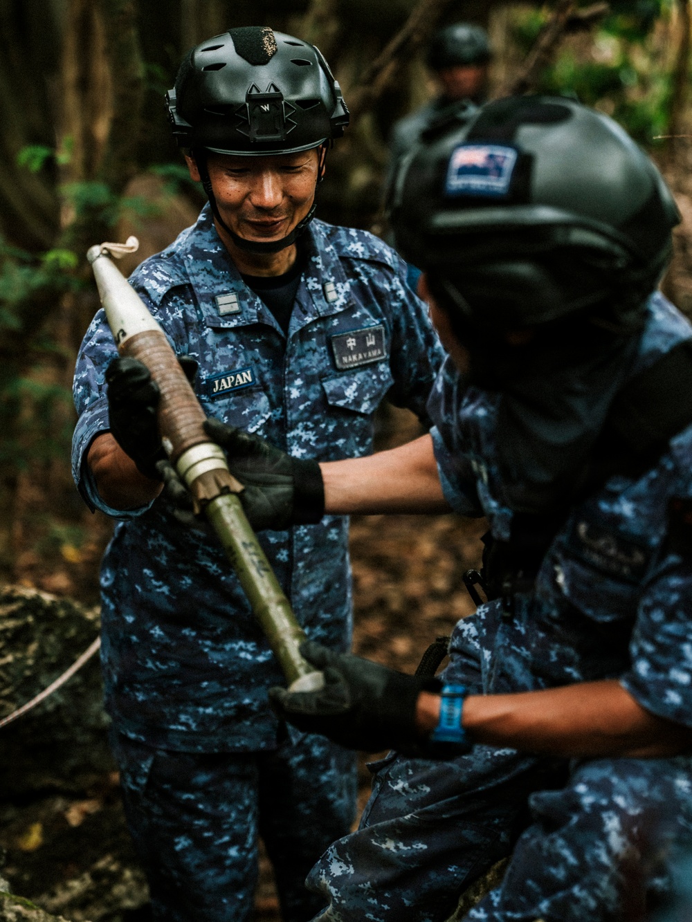 Two military personnel in Japanese Self-Defense Force blue digital camouflage uniforms are handling what appears to be an ordnance or demolition device in a forested outdoor setting. The standing individual is identified by name tape as 'NAKAYAMA' and wears a 'JAPAN' country patch. A third individual is partially visible in the background, and a second person wearing a helmet with what appears to be a New Zealand flag patch is visible in the foreground.