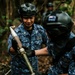 Two military personnel in Japanese Self-Defense Force blue digital camouflage uniforms are handling what appears to be an ordnance or demolition device in a forested outdoor setting. The standing individual is identified by name tape as 'NAKAYAMA' and wears a 'JAPAN' country patch. A third individual is partially visible in the background, and a second person wearing a helmet with what appears to be a New Zealand flag patch is visible in the foreground.
