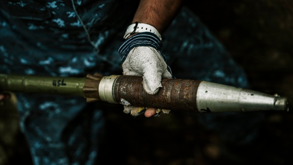 A gloved hand is holding what appears to be a rocket-propelled grenade (RPG) or similar munition. The object shows signs of age or wear, with rust and corrosion visible on the central body section. The person is wearing work gloves and appears to be handling the item carefully.