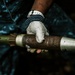 A gloved hand is holding what appears to be a rocket-propelled grenade (RPG) or similar munition. The object shows signs of age or wear, with rust and corrosion visible on the central body section. The person is wearing work gloves and appears to be handling the item carefully.