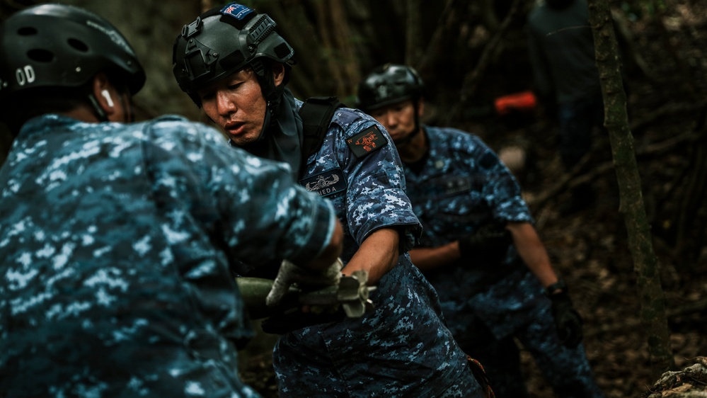 Military or law enforcement personnel in blue digital camouflage uniforms are engaged in a physical task in a forested outdoor environment. The central figure wears a tactical helmet with a flag patch and a name tag reading 'MEDA', along with a chest rig and unit patches. The personnel appear to be conducting a search, rescue, or debris-clearing operation.