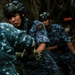 Military or law enforcement personnel in blue digital camouflage uniforms are engaged in a physical task in a forested outdoor environment. The central figure wears a tactical helmet with a flag patch and a name tag reading 'MEDA', along with a chest rig and unit patches. The personnel appear to be conducting a search, rescue, or debris-clearing operation.