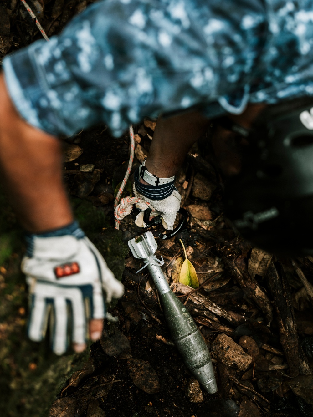 A person wearing military/digital camouflage uniform and protective work gloves is handling what appears to be an unexploded ordnance or mortar round on rocky ground covered with debris and leaves. The individual appears to be engaged in explosive ordnance disposal (EOD) or demining activity, as suggested by the protective gloves, uniform, and careful handling of the munition. A rope or cord is also visible near the ordnance.