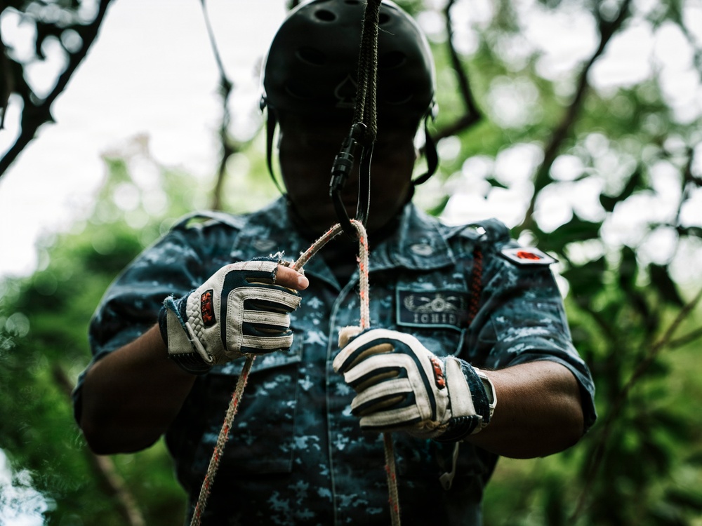 A person wearing military-style camouflage uniform and a black helmet is handling ropes and rigging equipment, likely preparing for a rappelling or zip-line activity. They are wearing white and black tactical gloves and appear to be in an outdoor, forested environment. The image is taken from a low angle, emphasizing the hands and rope work.