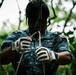 A person wearing military-style camouflage uniform and a black helmet is handling ropes and rigging equipment, likely preparing for a rappelling or zip-line activity. They are wearing white and black tactical gloves and appear to be in an outdoor, forested environment. The image is taken from a low angle, emphasizing the hands and rope work.