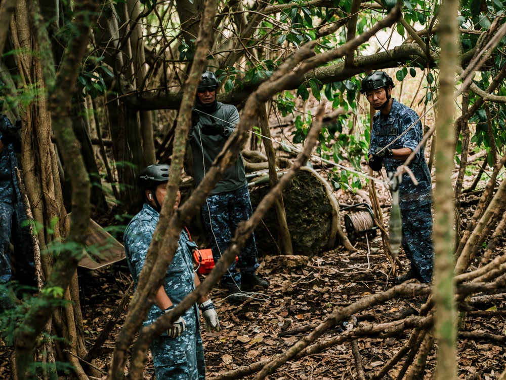 Several individuals wearing blue digital camouflage uniforms and helmets are working in a dense forested area. They appear to be handling ropes and equipment, suggesting a search, rescue, or military training operation in a jungle or woodland environment. The scene is partially obscured by tree branches and undergrowth.
