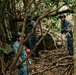Several individuals wearing blue digital camouflage uniforms and helmets are working in a dense forested area. They appear to be handling ropes and equipment, suggesting a search, rescue, or military training operation in a jungle or woodland environment. The scene is partially obscured by tree branches and undergrowth.
