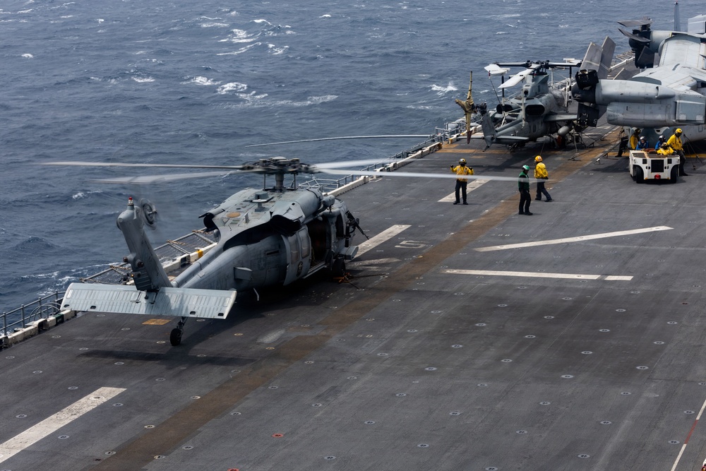 A military helicopter, appearing to be an MH-60 Seahawk, is positioned on the flight deck of a naval vessel at sea. Several deck crew members in yellow and green jerseys are visible performing flight deck operations. Additional rotary-wing aircraft, likely MV-22 Ospreys, are visible in the background along with a deck tractor.