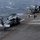 USS Boxer Conducts Flight Operations A military helicopter, appearing to be an MH-60 Seahawk, is positioned on the flight deck of a naval vessel at sea. Several deck crew members in yellow and green jerseys are visible performing flight deck operations. Additional rotary-wing aircraft, likely MV-22 Ospreys, are visible in the background along with a deck tractor.