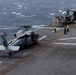 A military helicopter, appearing to be an MH-60 Seahawk, is positioned on the flight deck of a naval vessel at sea. Several deck crew members in yellow and green jerseys are visible performing flight deck operations. Additional rotary-wing aircraft, likely MV-22 Ospreys, are visible in the background along with a deck tractor.