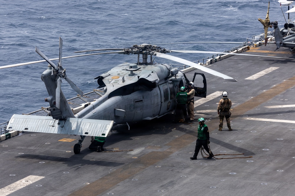 A military helicopter, identified as an MH-60 Seahawk variant, is secured on the flight deck of a naval vessel at sea. Several deck crew members in green jerseys and helmets are conducting pre-flight or post-flight operations around the aircraft. The ocean is visible beyond the ship's deck edge, indicating open-water operations.