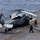 USS Boxer Conducts Flight Operations A military helicopter, identified as an MH-60 Seahawk variant, is secured on the flight deck of a naval vessel at sea. Several deck crew members in green jerseys and helmets are conducting pre-flight or post-flight operations around the aircraft. The ocean is visible beyond the ship's deck edge, indicating open-water operations.
