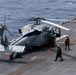 A military helicopter, identified as an MH-60 Seahawk variant, is secured on the flight deck of a naval vessel at sea. Several deck crew members in green jerseys and helmets are conducting pre-flight or post-flight operations around the aircraft. The ocean is visible beyond the ship's deck edge, indicating open-water operations.
