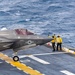 An F-35B stealth fighter jet from Marine Fighter Attack Squadron (VMFA) 122 is positioned on the flight deck of an amphibious assault ship at sea. The aircraft, numbered 118, is being directed by two flight deck crew members wearing yellow jerseys and helmets. The scene depicts pre-launch or taxiing operations with open ocean visible in the background.