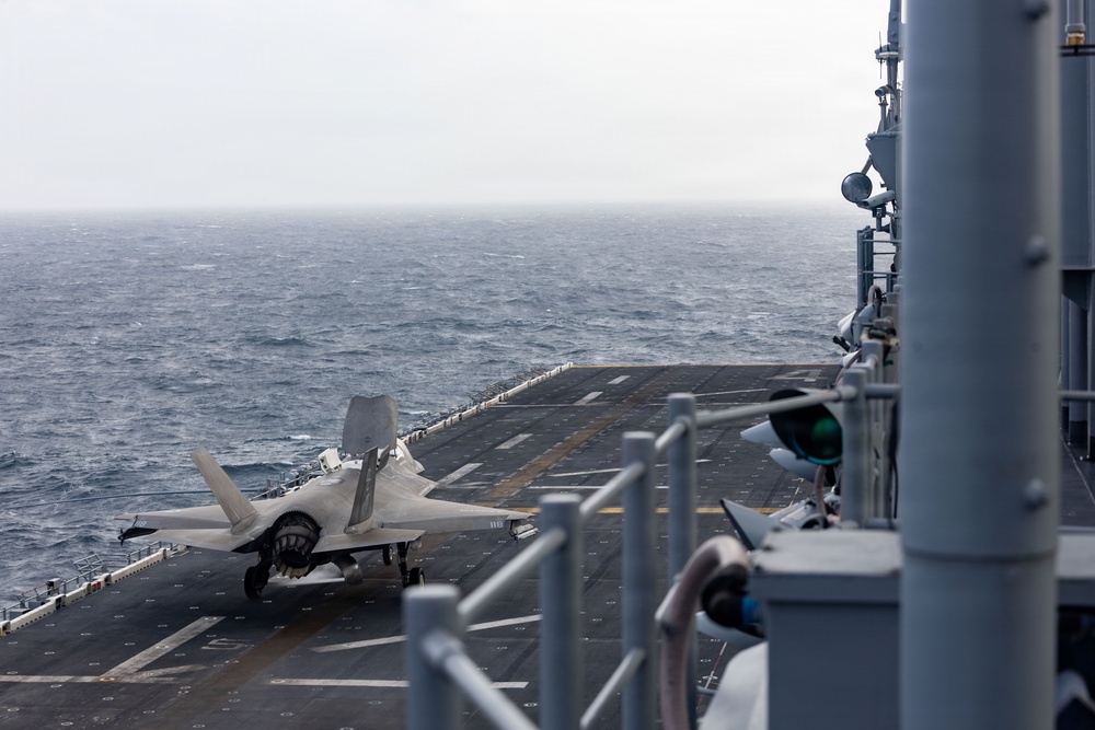 A stealth fighter jet, identifiable as an F-35B variant based on its distinctive shape and short takeoff/vertical landing configuration, is positioned on the flight deck of a naval vessel at sea. The aircraft appears stationary on the deck with its landing gear deployed. The image is taken from the ship's island superstructure looking down the flight deck toward the open ocean under overcast conditions.