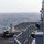 USS Boxer Conducts Flight Operations A stealth fighter jet, identifiable as an F-35B variant based on its distinctive shape and short takeoff/vertical landing configuration, is positioned on the flight deck of a naval vessel at sea. The aircraft appears stationary on the deck with its landing gear deployed. The image is taken from the ship's island superstructure looking down the flight deck toward the open ocean under overcast conditions.