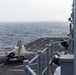 A stealth fighter jet, identifiable as an F-35B variant based on its distinctive shape and short takeoff/vertical landing configuration, is positioned on the flight deck of a naval vessel at sea. The aircraft appears stationary on the deck with its landing gear deployed. The image is taken from the ship's island superstructure looking down the flight deck toward the open ocean under overcast conditions.